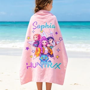 May include: A pink beach towel featuring a colorful graphic of four animated characters, a cat, and the text "HUNTRAX". The name "Sophia" is printed at the top. The towel is draped over a person on a sandy beach with the ocean in the background.