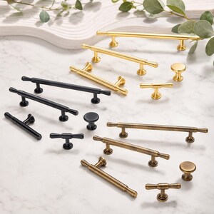 May include: A collection of cabinet hardware including various drawer pulls, T-bar handles, and round knobs. The hardware is available in three distinct finishes: polished gold, matte black, and an antique brass tone, featuring smooth cylindrical designs.