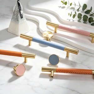 May include: A collection of cabinet hardware including three long bar pulls and three round knobs. The hardware features brushed gold metal ends with center sections wrapped in pink, blue, or orange leather. Some leather sections show stitching.