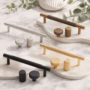 May include: A collection of cabinet hardware featuring T-bar handles and matching knobs in silver, antique brass, gold, and matte black finishes. Each piece displays a textured, knurled grip pattern on the cylindrical surfaces of the handles and knobs.