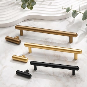 May include: A collection of cabinet hardware including three long cylindrical bar pulls and three matching T-shaped knobs. The set features three distinct finishes: a brushed antique brass, a bright satin gold, and a matte black metal finish.