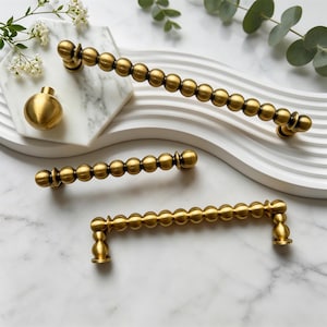 May include: A collection of brass-toned cabinet hardware including one round knob and three elongated drawer pulls. Each pull features a series of connected spheres along the handle, finished with a smooth, metallic sheen and matching mounting posts.