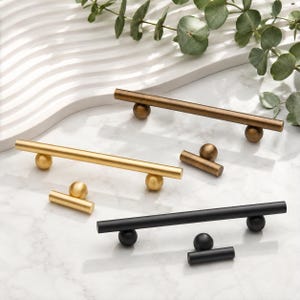 May include: A collection of cabinet hardware including three long horizontal bar pulls and three matching small T-shaped knobs. The hardware is finished in three distinct metallic colors: a dark bronze, a bright brushed gold, and a matte black finish.