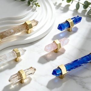 May include: A collection of cabinet knobs featuring double-pointed crystal-shaped bars in clear, smoky, pink, and blue hues. Each piece is secured by a central brushed gold-tone metal mount, showcasing a faceted geometric design.