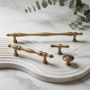 May include: A collection of four antique brass cabinet hardware pieces, including two elongated drawer pulls with a central bulge, one small T-shaped handle, and one round knob, all featuring a brushed metallic finish and rounded ends.
