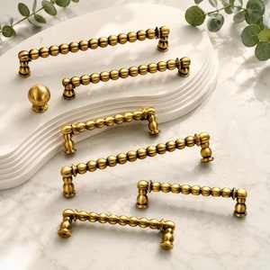 May include: A collection of brass-toned cabinet hardware including five elongated drawer pulls with a repeating spherical bead design and one matching round cabinet knob, all featuring a smooth metallic finish and sturdy mounting posts.