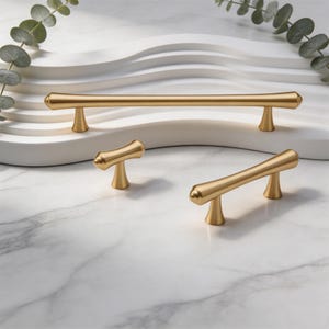 Puede incluir: Tres tiradores de armario y un pomo de metal en tono dorado con un acabado mate suave. Los herrajes presentan un diseño de barra cilíndrica larga y delgada con extremos cónicos y dos postes de montaje cónicos para cada pieza.