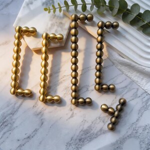 May include: A collection of metal cabinet hardware featuring a beaded design. The set includes two bright gold-toned rectangular handles, two darker bronze-toned rectangular handles, and one small bronze-toned T-shaped knob, all with a spherical texture.