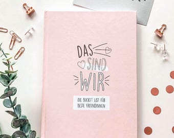 May include: A pink hardcover journal with the German text "DAS SIND WIR" and "DIE BUCKET LIST FÜR BESTE FREUNDINNEN". The journal is surrounded by stationery items, including paper clips, push pins, and a decorative sprig of greenery.