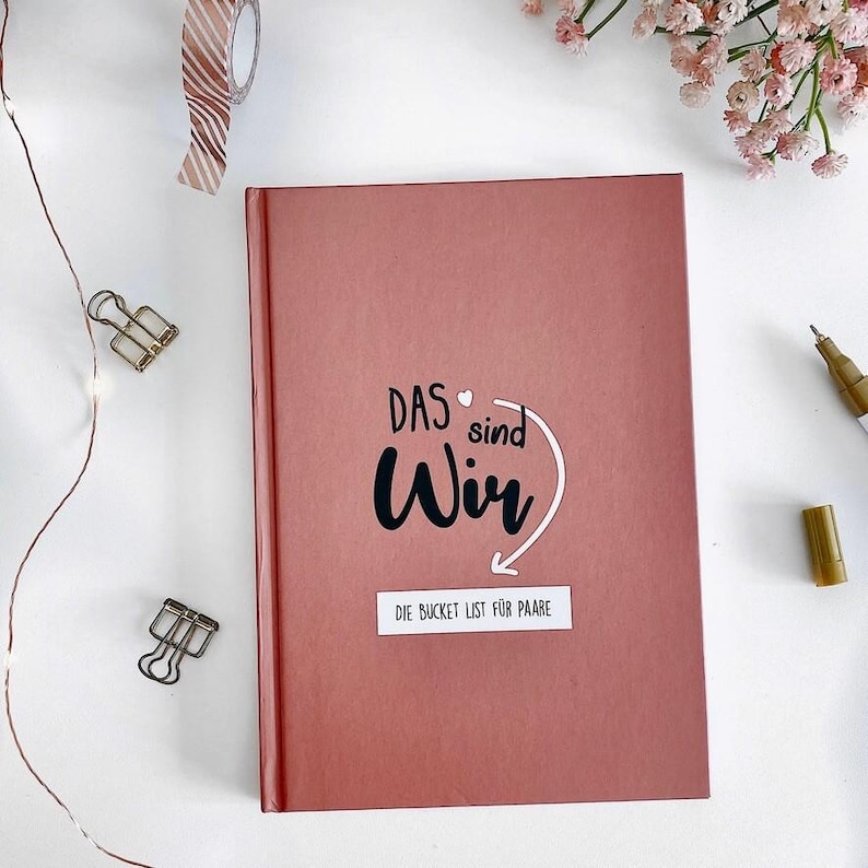 May include: A dusty rose-colored journal with the words "DAS sind Wir" and "DIE BUCKET LIST F&Uuml;R PAARE" in black and white. The journal is surrounded by decorative items, including a gold pen, paper clips, and washi tape.
