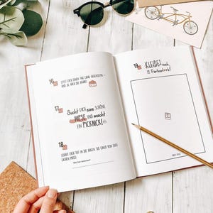 May include: An open journal with text in German, a pencil, and sunglasses on a white wooden surface. The journal pages have pink edges and include text and illustrations. A small bicycle illustration is on a card.