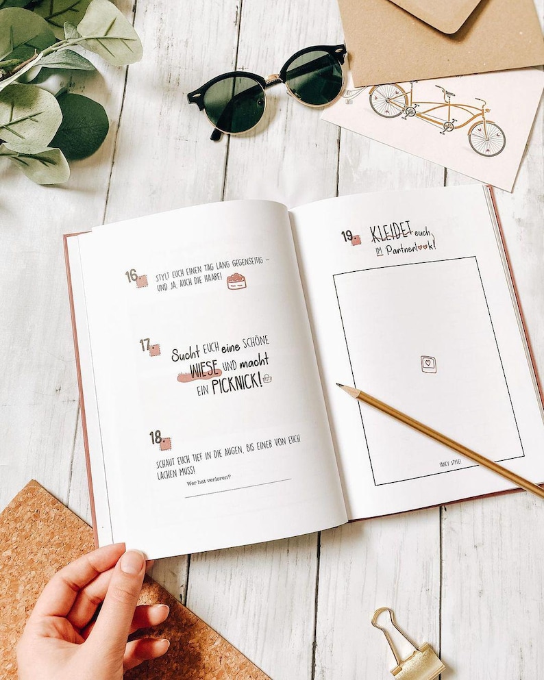 May include: An open journal with text in German, a pencil, and sunglasses on a white wooden surface. The journal pages have pink edges and include text and illustrations. A small bicycle illustration is on a card.
