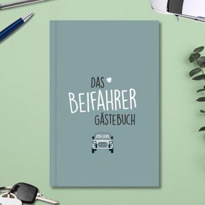 Könnte beinhalten: Ein türkisblaues Gästebuch mit dem deutschen Text "DAS BEIFAHRER GÄSTEBUCH" und einer kleinen Autoillustration. Das Buch ist geschlossen und hat ein schlichtes Design. Ein Stift, Autoschlüssel und ein Spielzeugauto sind sichtbar.