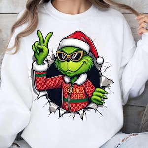 Może przedstawiać: Biała bluza z zieloną postacią Grincha w czapce Świętego Mikołaja i okularach przeciwsłonecznych, pokazującą znak pokoju. Grinch ma na sobie czerwoną kurtkę z wzorem w stylu Gucci, wychodzącą z podartej dziury.