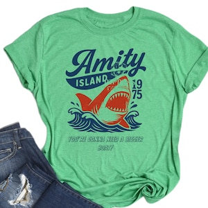 May include: Green t-shirt with a graphic featuring a shark leaping from ocean waves. The design includes the words "Amity Island 1975" and the phrase "You're Gonna Need a Bigger Boat!"