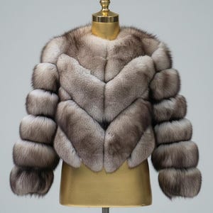 May include: A luxurious, short fur jacket in shades of gray and brown, displayed on a golden mannequin. The jacket features a chevron design on the front and unique, layered sleeves. The overall style is elegant and sophisticated.