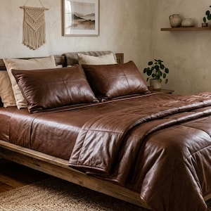 Op de afbeelding: Een bed met een bruin leren dekbed en bijpassende kussens. Het bedframe is van hout. Een klein houten nachtkastje met een lamp en boeken staat naast het bed. De kamer heeft een neutraal kleurenpalet.