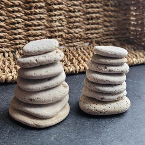 May include: Two stacks of smooth, light gray stones, each stacked in a balanced tower. The stones are arranged on a dark gray surface.