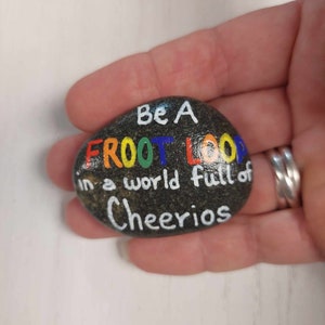 May include: A painted rock with the words "Be a Froot Loop in a world full of Cheerios" in rainbow colors.