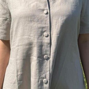 以下が含まれることがあります： 丸いネックラインと前面中央に縦一列に並んだ共布のボタンが特徴のライトベージュの半袖ドレス。襟元付近には対照的な2つのダークカラーのボタンが見えます。