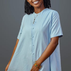 Puede incluir: Vestido abotonado azul claro con mangas cortas. El vestido llega a la rodilla y tiene un corte holgado. Se ven un collar dorado y una pulsera de cuentas. El fondo es gris liso.