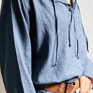 May include: A blue tunic with long sleeves and a V-neck, tied with drawstrings. A brown leather belt with a buckle is worn at the waist. A beaded necklace is around the neck. The clothing has a medieval or renaissance style.