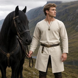 May include: A beige long-sleeved tunic with a round neckline and a centre front slit, worn with a dark brown leather belt featuring a decorative buckle and a pendant necklace, standing beside a black horse with a leather bridle and harness.