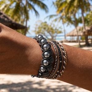 May include: A multi-strand bracelet featuring a row of smooth, metallic gray spherical beads interspersed with dark, porous textured beads, alongside several braided cords in shades of black, tan, and white, wrapped around a wrist.