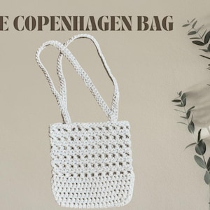 May include: A white, crocheted bag with two handles, labeled "THE COPENHAGEN BAG" in brown text. The bag has a rectangular shape with a textured, open weave design. A sprig of green leaves is in the background.