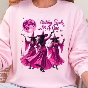 May include: A light pink sweatshirt featuring a graphic of three witches in magenta dresses and hats, with a full moon and the text "Casting Spels for A Cure". Butterflies and candles are also included in the design.