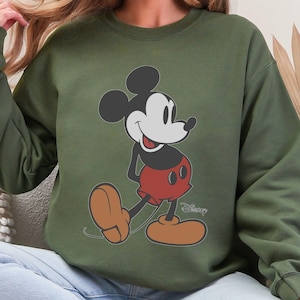 May include: Olive green sweatshirt featuring a classic Mickey Mouse graphic. The cartoon character is depicted in his iconic pose, with black ears, a white face, red shorts, and brown shoes. The Disney logo is printed below the character.
