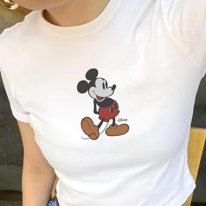 Puede incluir: Camiseta blanca con un gr&aacute;fico de Mickey Mouse. El personaje de dibujos animados se muestra con pantalones cortos rojos, zapatos marrones y orejas negras. El logotipo de Disney es visible en la parte inferior derecha.