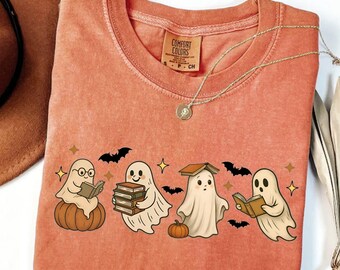 May include: Coral-colored long-sleeve shirt featuring a graphic of four cartoon ghosts reading books, with pumpkins, bats, and stars. The shirt has a crew neck and the Comfort Colors brand tag. The word "YAM" is printed at the bottom left.