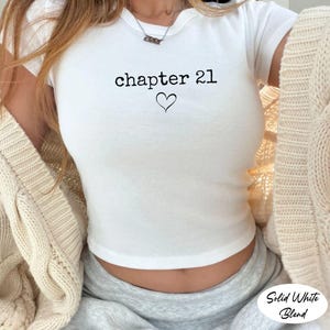 May include: White crop top with the text "chapter 21" and a heart graphic. The shirt is a solid white color and has short sleeves. The words "Solid White Blend" are in the bottom right corner.