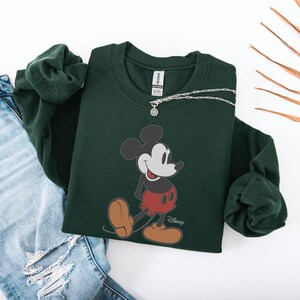 May include: A dark green sweatshirt featuring a graphic of Mickey Mouse in his classic pose. Mickey is depicted in black, white, red, and tan. A silver necklace with a small pendant rests on the sweatshirt. The sweatshirt is paired with blue jeans.