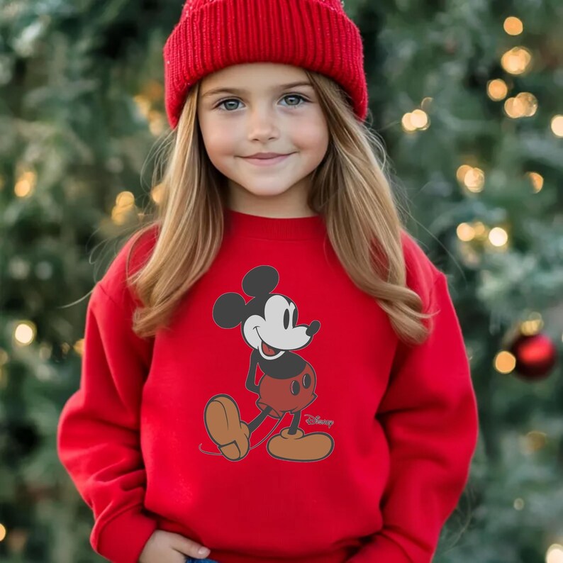 May include: A red sweatshirt featuring a Mickey Mouse graphic. The sweatshirt is paired with a matching red beanie. The design includes a black and white Mickey Mouse illustration with red shorts and brown shoes. The background is a blurred Christmas tree.
