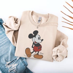 May include: Beige sweatshirt featuring a classic Mickey Mouse graphic. The cartoon character is depicted in black, red, and brown, with the Disney logo below. The sweatshirt is paired with blue jeans and a delicate silver necklace.