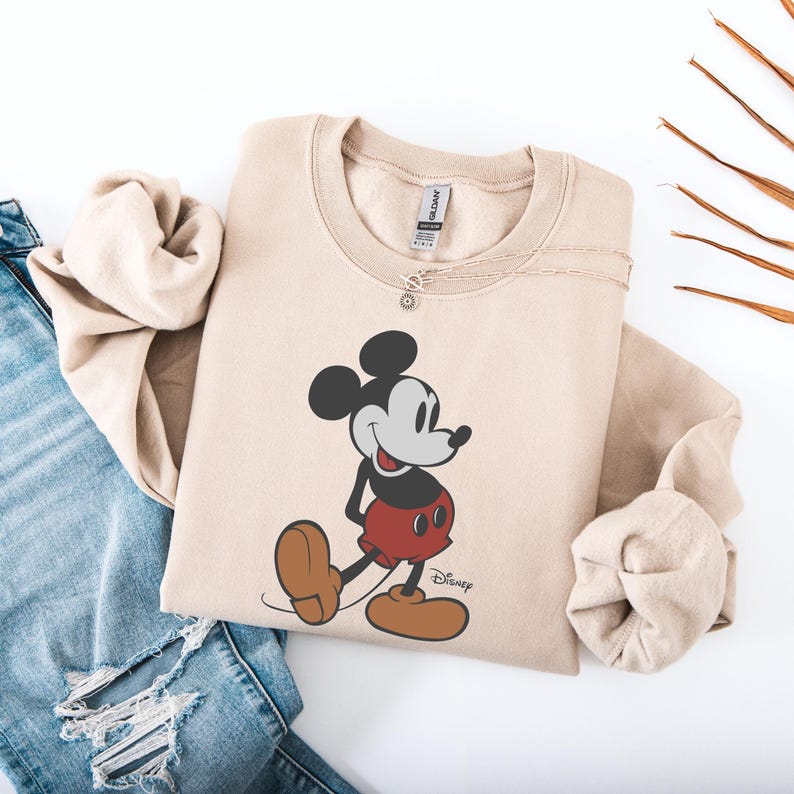 May include: Beige sweatshirt featuring a classic Mickey Mouse graphic. The cartoon character is depicted in black, red, and brown, with the Disney logo below. The sweatshirt is paired with blue jeans and a delicate silver necklace.