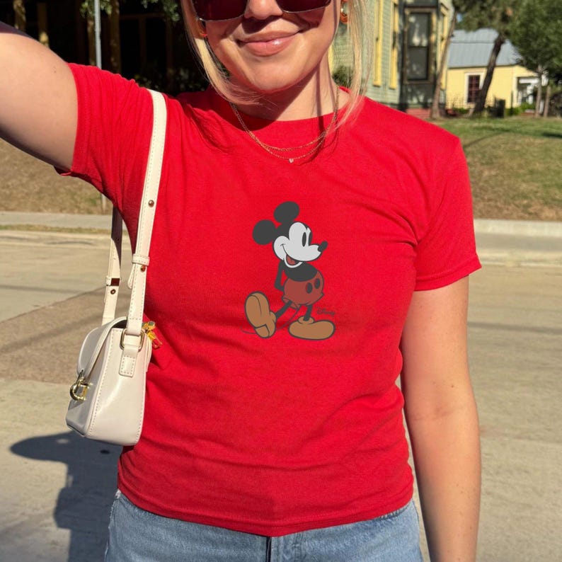 Puede incluir: Camiseta roja con un dibujo de Mickey Mouse. La camiseta tiene mangas cortas y cuello redondo. Se ve un bolso blanco roto con una correa para el hombro.