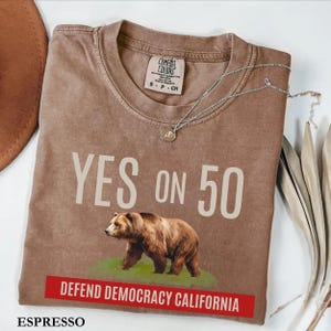May include: Espresso-colored t-shirt with the text "YES ON 50" above a brown bear graphic. Below the bear is a red banner that reads "DEFEND DEMOCRACY CALIFORNIA." A silver necklace with a pendant is draped across the shirt.