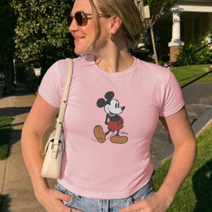 Puede incluir: Camiseta rosa claro con un dibujo de Mickey Mouse. El personaje de dibujos animados est&aacute; representado con su atuendo cl&aacute;sico: pantalones cortos rojos, zapatos marrones y guantes blancos. La camiseta tiene mangas cortas y cuello redondo.