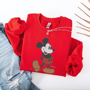 May include: A red sweatshirt featuring a graphic of Mickey Mouse. The cartoon character is depicted in black, white, and red, with brown shoes. A silver necklace is draped across the sweatshirt. The sweatshirt is paired with blue jeans.