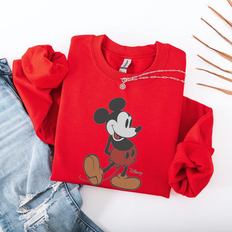 May include: A red sweatshirt featuring a graphic of Mickey Mouse. The cartoon character is depicted in black, white, and red, with brown shoes. A silver necklace is draped across the sweatshirt. The sweatshirt is paired with blue jeans.