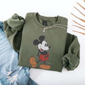 May include: An olive green sweatshirt featuring a graphic of Mickey Mouse in his classic pose. The sweatshirt has a crew neck and long sleeves. A silver necklace is draped across the top. The sweatshirt is paired with a pair of blue jeans.