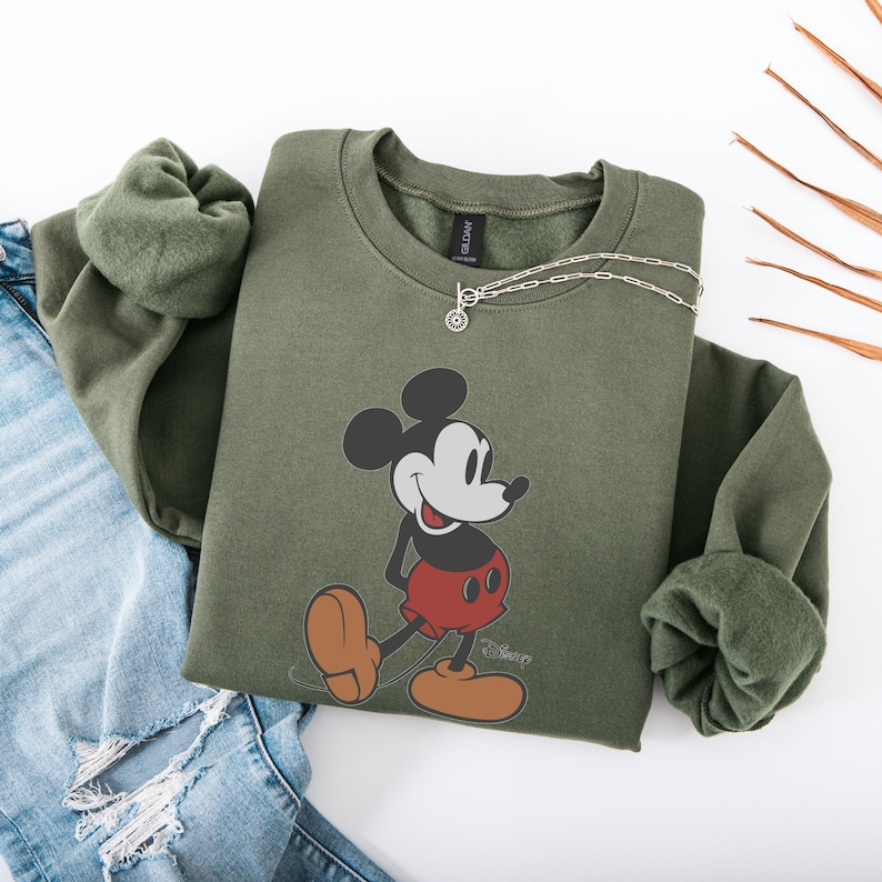 May include: An olive green sweatshirt featuring a graphic of Mickey Mouse in his classic pose. The sweatshirt has a crew neck and long sleeves. A silver necklace is draped across the top. The sweatshirt is paired with a pair of blue jeans.