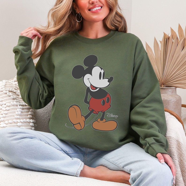 May include: Olive green sweatshirt featuring a classic Mickey Mouse graphic. The cartoon character is depicted in his iconic pose, with black ears, a white face, red shorts, and brown shoes. The Disney logo is printed below the character.