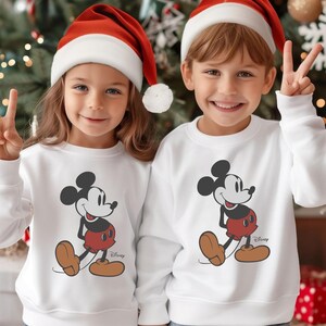 May include: Two children wearing white sweatshirts featuring a Mickey Mouse graphic, red shorts, and brown shoes. Both children are wearing red and white Santa hats and smiling. The Disney logo is visible on the sweatshirts.