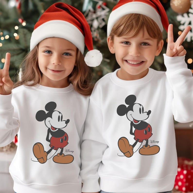 May include: Two children wearing white sweatshirts featuring a Mickey Mouse graphic, red shorts, and brown shoes. Both children are wearing red and white Santa hats and smiling. The Disney logo is visible on the sweatshirts.