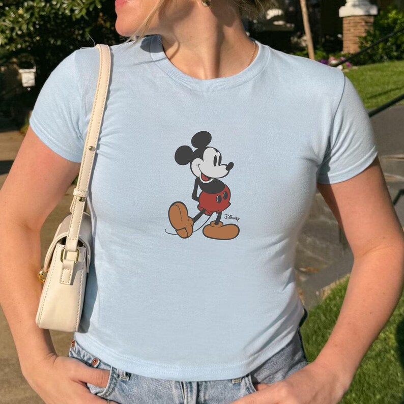 Puede incluir: Camiseta azul claro con un dise&ntilde;o de Mickey Mouse. El personaje de dibujos animados se muestra en su pose cl&aacute;sica, con pantalones cortos rojos y zapatos marrones. El logotipo de Disney es visible en la parte inferior derecha.