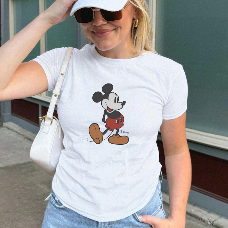 Puede incluir: Camiseta blanca con un gr&aacute;fico de Mickey Mouse. El personaje de dibujos animados est&aacute; representado en negro, rojo y marr&oacute;n, con el logotipo de Disney debajo. La camiseta es de estilo casual.